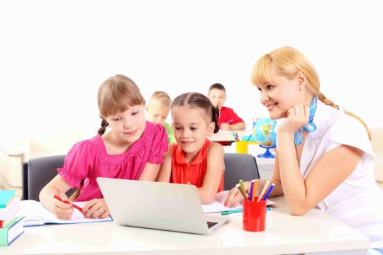 Female teacher sitting at a table with young students hovering over a laptop