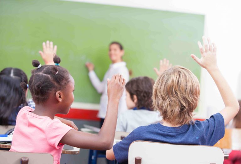 Young students sitting at their desks in a classroom with their hands raised