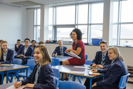 Laughing female teacher sitting with smiling students in a classroom.