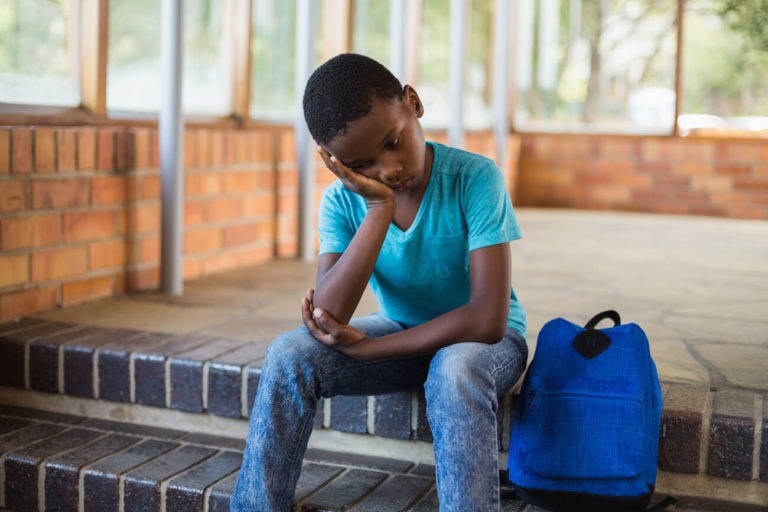 Sad boy sitting with his head in his hand on school steps with his backpack.