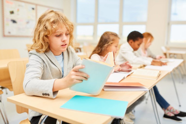 Young boy reading a book at his desk in a classroom with other students.