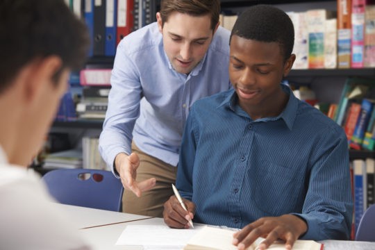 Male student working at a table with male teacher helping.