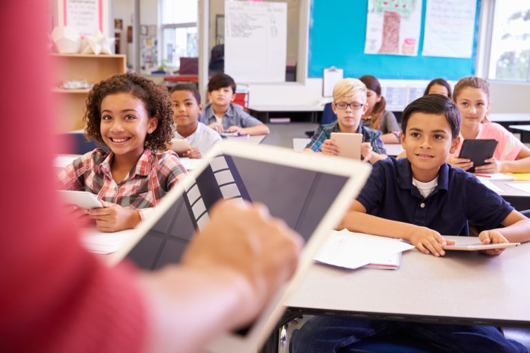 Teacher standing in the front of a classroom holding a tablet with students at their desks with tablets.