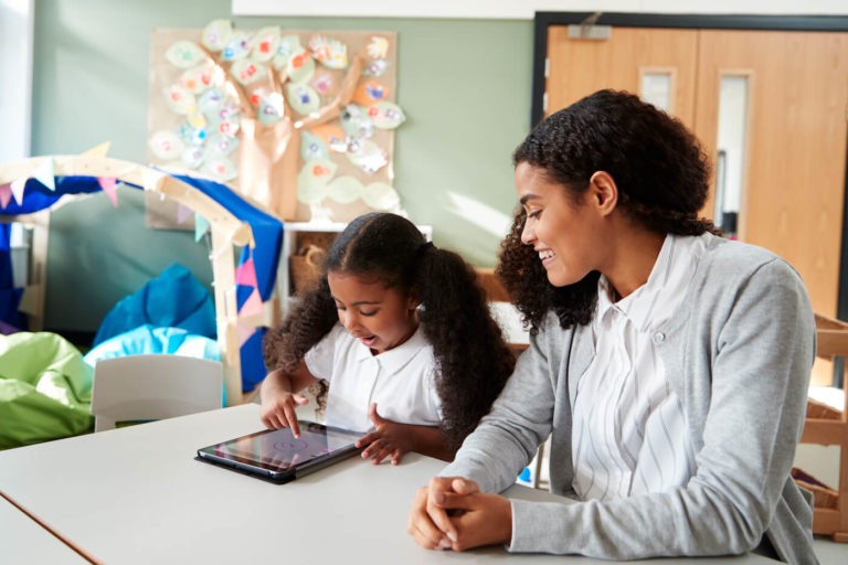 Young girl sitting at a table and using a tablet next to a teacher.