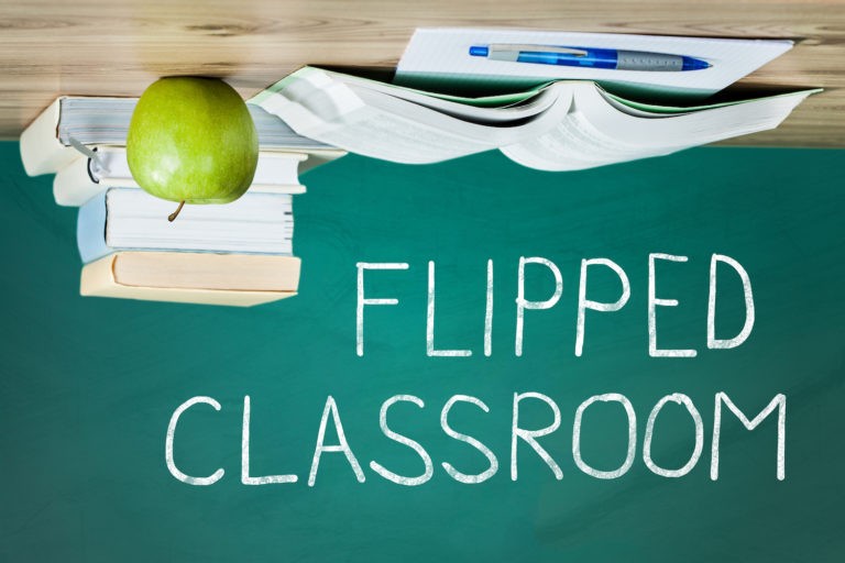 A table with books, an apple, and a pen flipped upside down with the words ‘flipped classroom’ written in chalk.