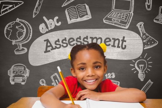 Young, smiling girl sitting at a desk with a pencil and paper with assessment written on the blackboard behind her.