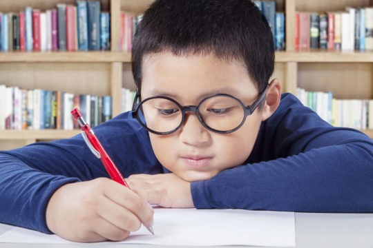 Young boy sitting at a table with head down writing on paper