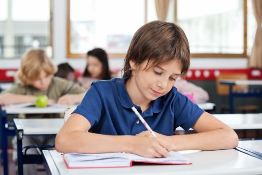 Young boy sitting at a desk in a classroom writing in a journal.