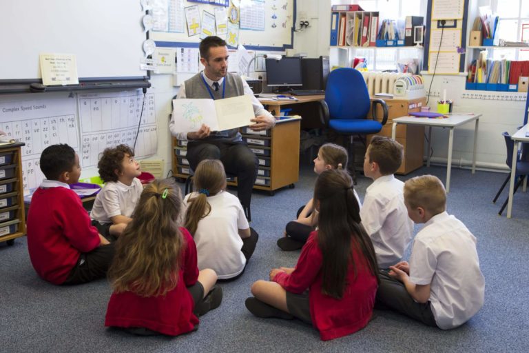 Group of children sitting on a classroom floor while a teacher reads a book to them.