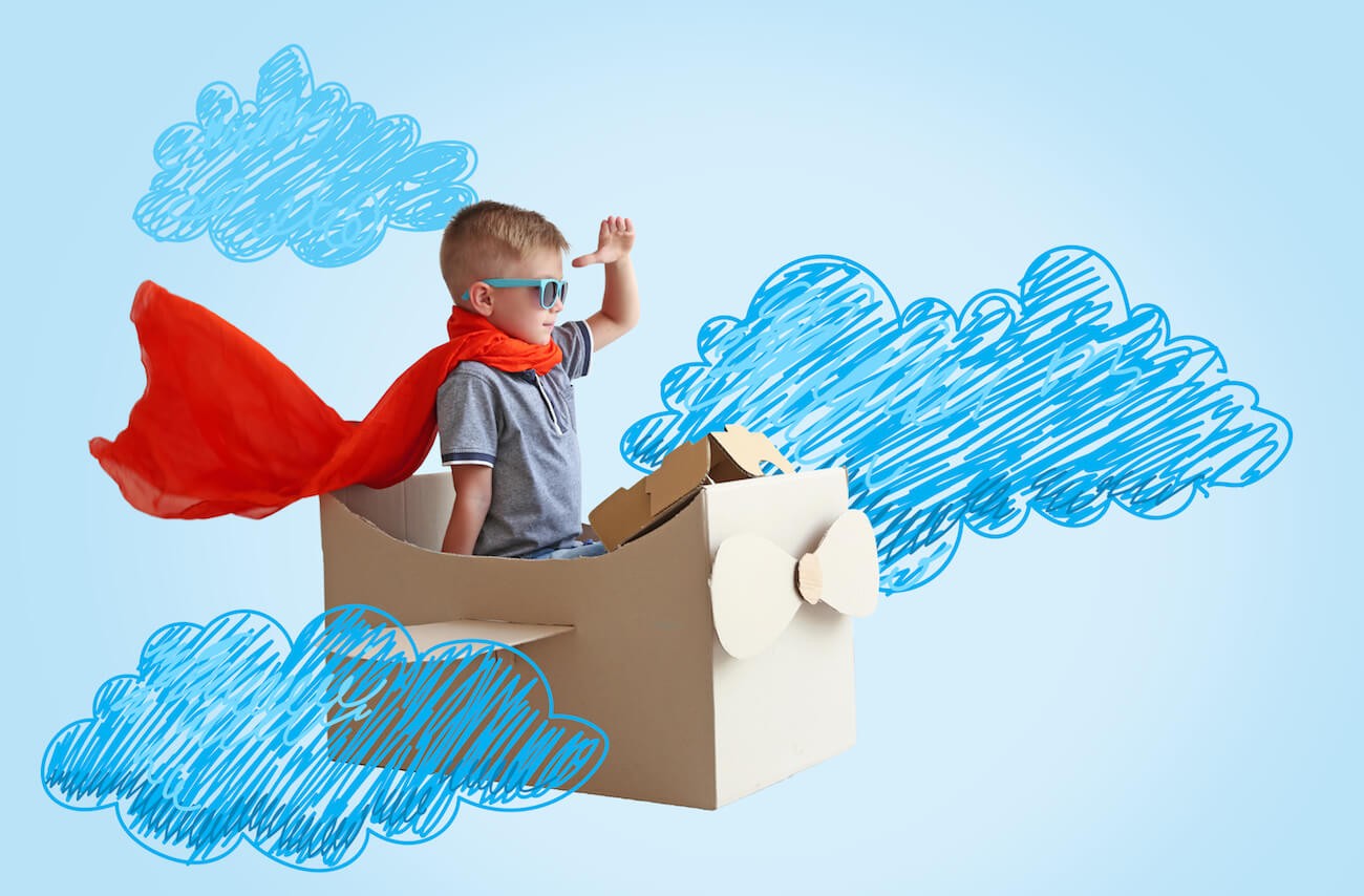 Young boy wearing a cape sitting in card board box pretending that he’s flying