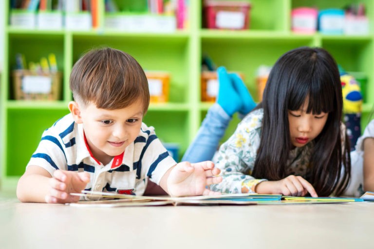 Two young students reading books on the ground in a classroom.