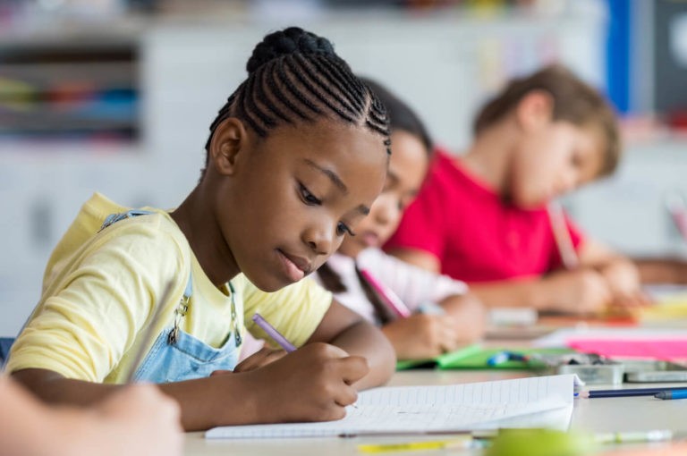 Young girl writing in a notebook among other students in a classroom.