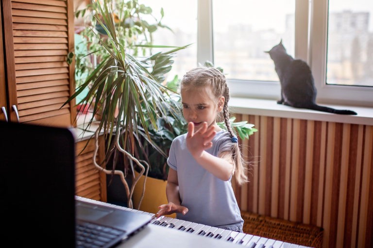 Young girl standing playing piano and waving at a laptop.