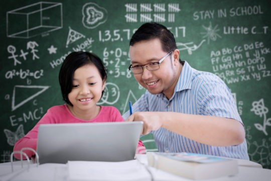 Student and teacher sitting at a computer with math equations written on chalkboard behind them