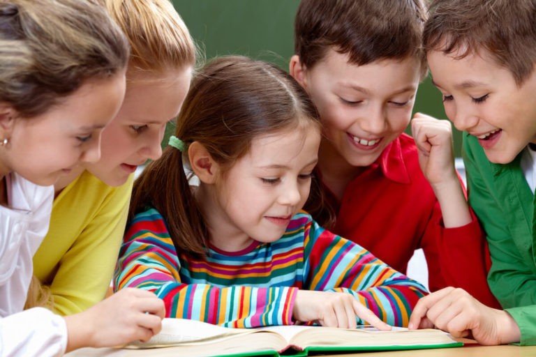 Group of young students smiling while reading a book together in a classroom.