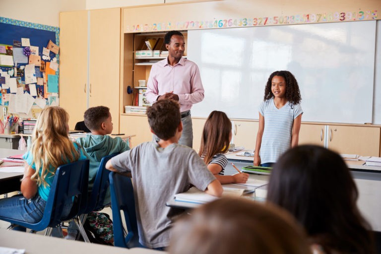 Young girl standing at the front of her elementary classroom with her teacher.