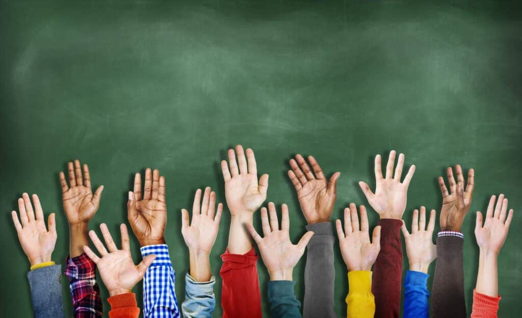A close-up of student hands being raised in front of a chalkboard, representing student participation.