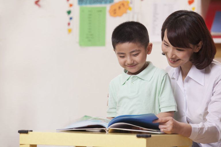 Smiling teacher helping young boy read sitting at a desk.