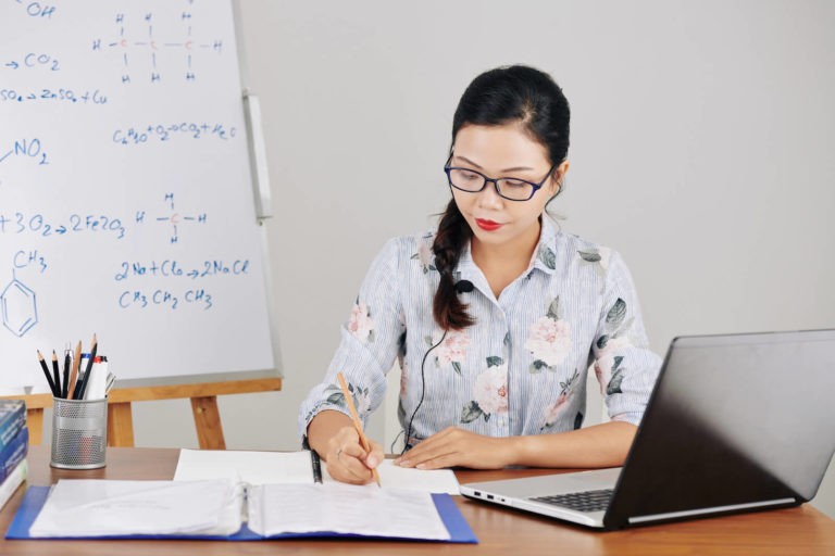 Teacher sitting at her desk taking notes and using a laptop.