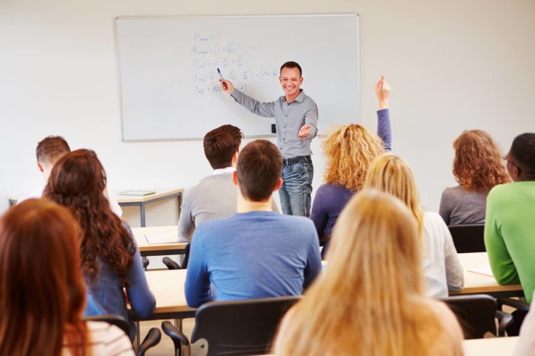 Smiling teacher pointing the a whiteboard and calling on a students with her hand up.