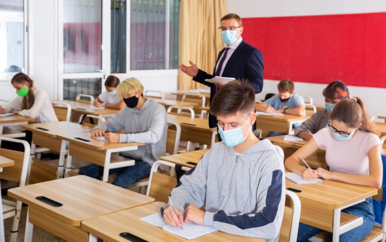 Teacher walking around a classroom while students work at their desks, all wearing masks.