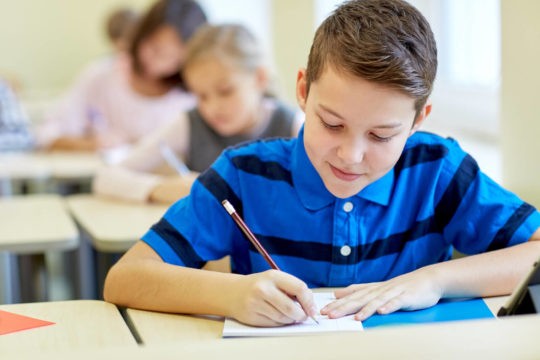 Content looking boy sitting at his desk writing in a classroom with students.