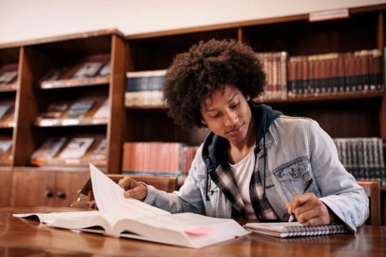 High school student sitting at a table in a library taking notes while reading a book.