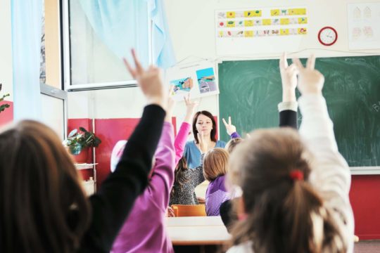 Students in a classroom raising two fingers in the air with a teacher standing in the front.