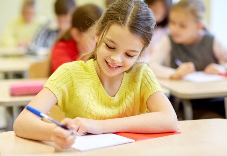 Young girl sitting at a desk in a classroom writing on paper.