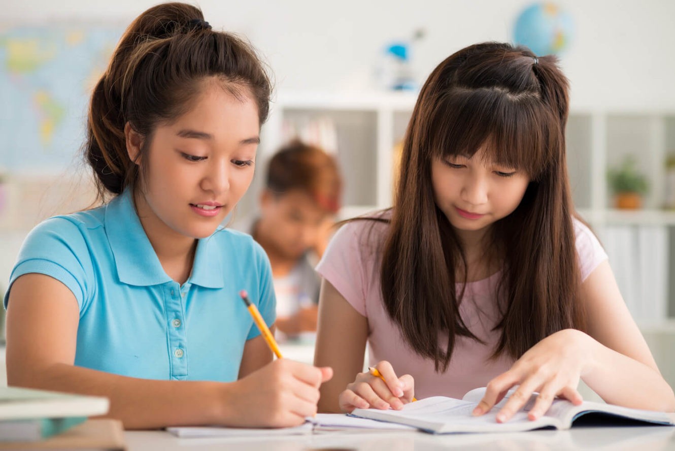 Two girls working together writing and reading at their desks