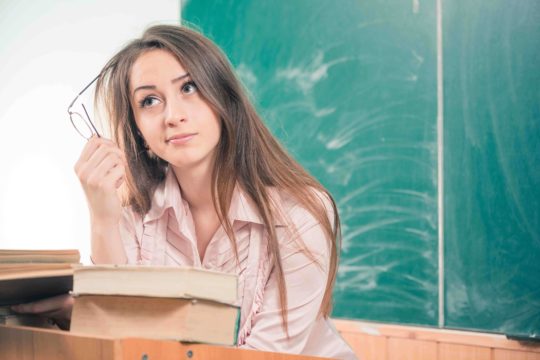 Young female teacher sitting in front of a chalkboard holding a pair of glasses while deep in thought