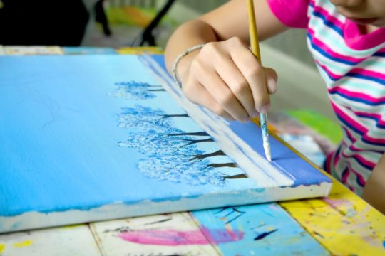 A young girl sitting at a table painting a winter scene on canvas.