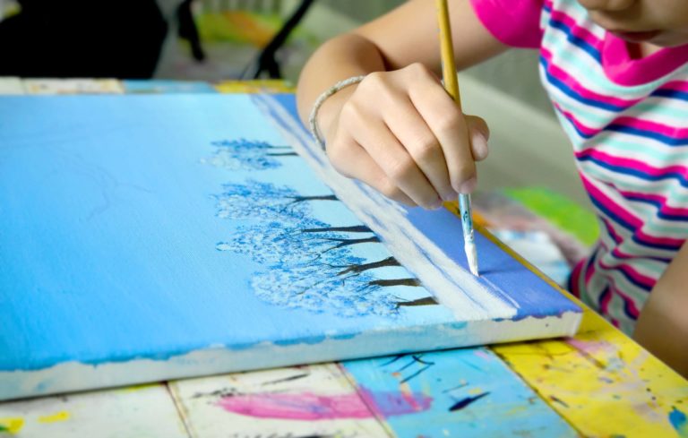 A young girl sitting at a table painting a winter scene on canvas.