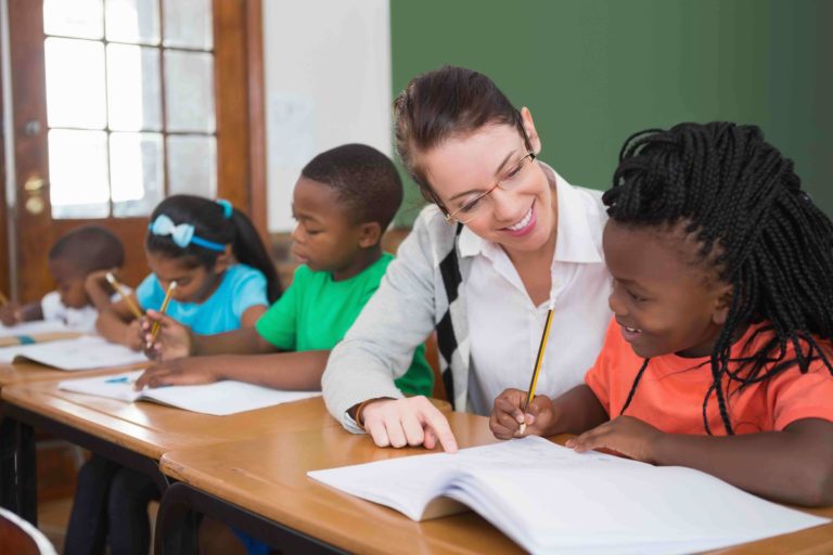 Female teacher helping a student writing in a notebook at their desk.